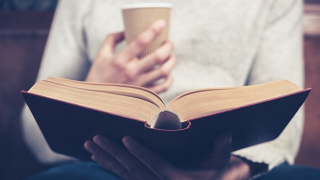 Sicht auf ein aufgeschlagenes Buch und einen Kaffeebecher, die ein Mensch in den Händen hält